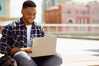 Başarılı bir Freelancer. Mutlu Afrikalı Adam gülümsüyor ve bilgisayarla çalışıyor, halka açık bir parkta oturuyor.
