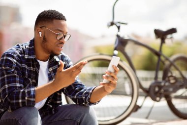 Siyah hipster 'ın cep telefonuyla video görüşmesi yaptığı, parkta oturduğu, kulaklık ve gözlük taktığı portresi.