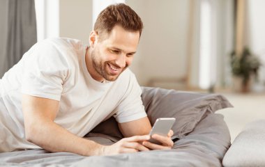 Mobil Uygulama. Evdeki yatak odasında yatarken akıllı telefon kullanan neşeli bir adam var. Telefondaki interneti tarayan adam. Cep telefonu reklamı için başvuru formu. Metin için Boş Alan