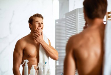 Erkek yüz derisi rutin konsepti. Tıraşlı, genç, yakışıklı bir adam sabahları tıraş olduktan sonra pürüzsüz çenesine dokunuyor, modern banyoda aynaya bakıyor..