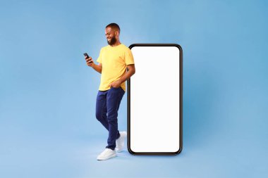 Yakışıklı Afrikalı genç bir Amerikalı cep telefonu kullanıyor. Mavi stüdyo arka planında boş ekranı olan büyük bir akıllı telefonun yanında duruyor. Web sitesi ya da uygulama için model, reklam için alan