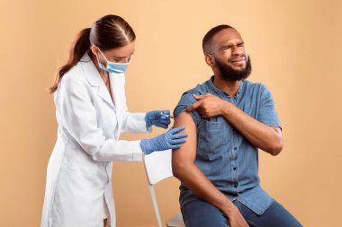 Genç Afrikalı Amerikalı gözlerini kapatıyor, grip aşısı sırasında bej rengi stüdyo geçmişinden korkuyor. Bayan doktor ya da hemşire hasta adama grip aşısı yapıyor.