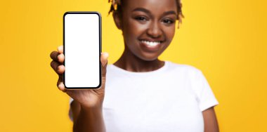 Mobil uygulama kavramı. Neşeli siyah kadın sarı arka planda boş ekranlı akıllı telefon gösteriyor. Beyaz ekran ile gülümseyen ve cep telefonu tutan Afrikalı Amerikalı genç bayana yakın çekim