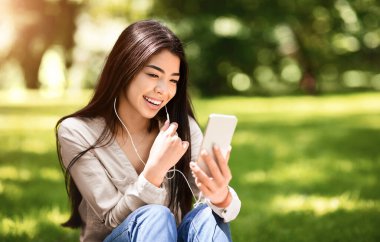 Genç Asyalı kız parkta dinlenirken akıllı telefondan video görüşmesi yapıyor. Yazın şehirde eğleniyor, boş yer var.
