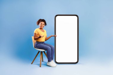 Mobil uygulama ya da web sitesi tasarımı için tasarlanmış, mavi stüdyo arka planı olan boş akıllı telefonun yanında oturan Afrikalı güzel bir bayan. Çevrimiçi reklamınız için boşluk