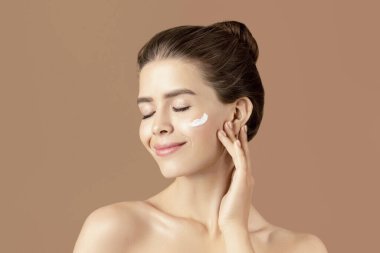 Güzellik ve sağlık konsepti. Kahverengi stüdyo arka planına yüz kremi süren hoş bir genç kadın. Milenyum hanımı cildine iyi bakıyor, nemlendiriyor ve besliyor.