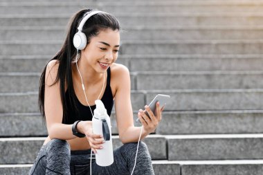 Eğitimden sonra. Sportif Asyalı kız merdivenlerde oturuyor, akıllı telefondan müzik dinliyor, bir şişe suyla antrenmandan sonra dinleniyor, boşluğu kopyalıyor.