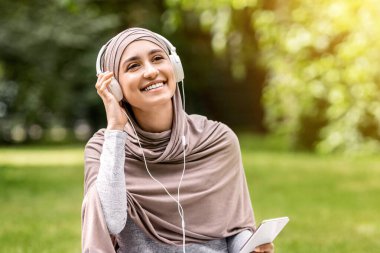 Başörtülü mutlu kadın parkta dinlenirken müziği seviyor, kulaklık ve akıllı telefon kullanıyor, boşluğu kopyalıyor