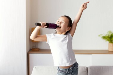 Küçük Singer. Sevimli bir kız şarkı söylüyor mikrofon gibi saç fırçası tutuyor evde koltukta dururken eğleniyor.