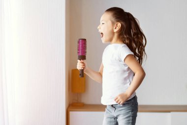 Küçük kız kanepede zıplayıp evde mikrofon gibi saç fırçası tutarak eğleniyor. Childs Boş Zaman Konsepti.