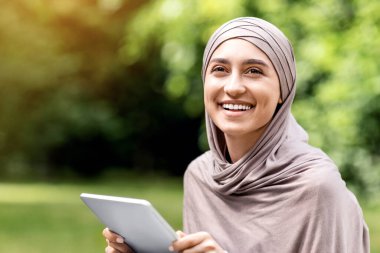Dijital tablet kullanan, halka açık parkta dinlenen mutlu Müslüman kadın.