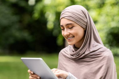 Tesettürde gülümseyen kız dijital tablet kullanıyor, parkta tek başına dinleniyor, boş bir alan.