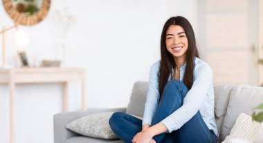 Sıcak bir hafta sonu. Evdeki rahat koltukta oturan, poz veren ve kameraya bakan, içtenlikle gülümseyen, şık bir oturma odasında dinlenen, izin gününün keyfini çıkaran mutlu Asyalı bir kadın.