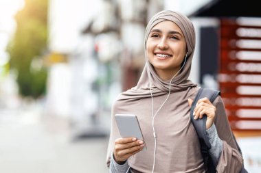 Caddede yürüyen, müzik dinleyen, akıllı telefon kullanan ve kulaklık takan mutlu Müslüman kadın.