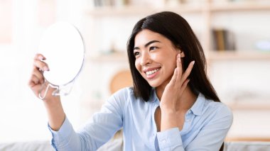 Doğal Güzellik. Güzel Asyalı kız evde büyüteç aynasına bakıyor ve kusursuz yumuşak tenine dokunuyor, yakın plan.