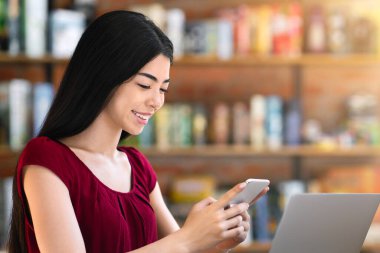 Neşeli Asyalı bayan girişimci, kafede dizüstü bilgisayarla çalışırken akıllı telefon kullanıyor, masada oturuyor ve kahve içiyor.