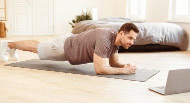 Sportif adam laptopta antrenman yapıyor, evde plank egzersizi yapıyor. Sabahları antrenman yapan adam spor rutini yapıyor. Sportif Yaşam Tarzı, Erkek Kilosu Kaybı Konsepti. Panorama