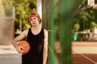 Spor giyiminde yakışıklı bir milenyum oyuncusu açık hava basketbol sahasında oynamaya hazır, boş bir alanda. Genç sporcu kameraya bakıyor ve maçtan sonra poz veriyor.