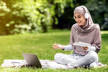 Aab kızı halka açık parkta ders çalışıyor, dizüstü bilgisayar ve kulaklık kullanıyor, notlar alıyor, yer kopyalıyor.