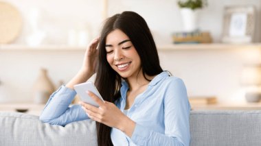 Rahatlatıcı bir hafta sonu. Mutlu Koreli kadın koltukta cep telefonuyla dinleniyor, sosyal ağları kontrol ediyor, arkadaşlarıyla mesajlaşıyor, tatilin tadını çıkarıyor, internetten alışveriş yapıyor, oturma odasında koltukta oturuyor, boşluğu kopyalıyor.