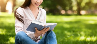 Gülümseyen Asyalı genç kız dışarıda kitap okuyor, parkta çimlerde dinleniyor, güneşli günün tadını çıkarıyor ya da sınava hazırlanıyor.