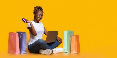 E-ticaret konsepti. Çevrimiçi alışveriş yapan pozitif siyah kız, dizüstü bilgisayar ve kredi kartı kullanıyor, alışveriş torbalarıyla yerde oturuyor, sarı arka plan, kopyalama alanı