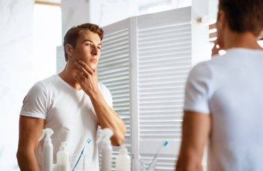 Ciddi yakışıklı bir adam yüzüne dokunuyor, banyodaki aynaya bakıyor.