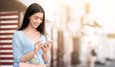 Çevrimiçi bankacılık. Genç Asyalı kadın alışveriş yaptıktan sonra kredi kartı bakiyesini akıllı telefonla kontrol ediyor, dışarıda alışveriş torbalarıyla dikiliyor.