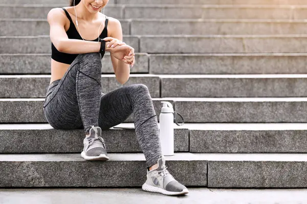 Kentsel Spor Konsepti. Gülümseyen Asyalı kız dışarıda koşmadan önce Smartwatch ayarlarını kontrol ediyor beton merdivenlerde bir şişe suyla oturuyor.