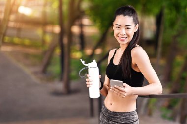 Koreli kız dışarıda antrenman yaptıktan sonra dinleniyor, su içiyor, akıllı telefon kullanıyor, kameraya gülümsüyor, boş alan arıyor.