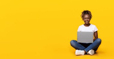 Siyah neşeli kız yerde oturuyor, dizüstü bilgisayar kullanıyor, sarı stüdyo arka planı, kopyalama alanı