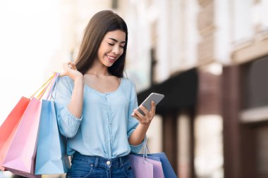 Güzel Asyalı kadın akıllı telefonunu açık havada alışveriş torbalarıyla dolaşırken kullanıyor.