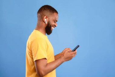 Bin yıllık Afro-Amerikan bir adamın akıllı telefon kullanışının mavi stüdyo arka planında kulaklıkla iletişim kurmasının, kopyalama alanının yan görüntüsü. Havalı adam mobil cihazda sanal toplantı yapıyor.