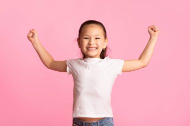 Zafer Konsepti. Elleri havada neşeli Çinli bir kız kazanan jestler yapıyor, pembe arka plan.
