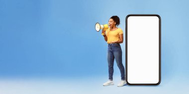 Mutlu genç Afrikalı Amerikalı kadın megafona bağırıyor, dev akıllı telefonun yanında boş ekranla duruyor, cep telefonu uygulamanızı veya web sitenizi mavi arka planda tanıtıyor, tasarım için modelleme yapıyor.