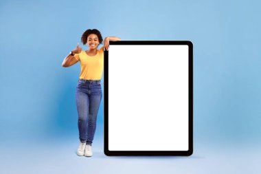 Büyük bir tablet bilgisayarın yanında boş ekranla duran mutlu Afro-Amerikalı kadın, başparmağını kaldırıyor, yeni uygulama ya da web sitesi öneriyor, çevrimiçi reklamınız için modelleme yapıyor, mavi arka plan.