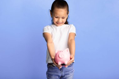 Bu benim param. Heyecanlı Asyalı çocuk pembe domuz kumbarası tutuyor ve ona bakıyor, mavi stüdyo duvarı, kopyalama alanı