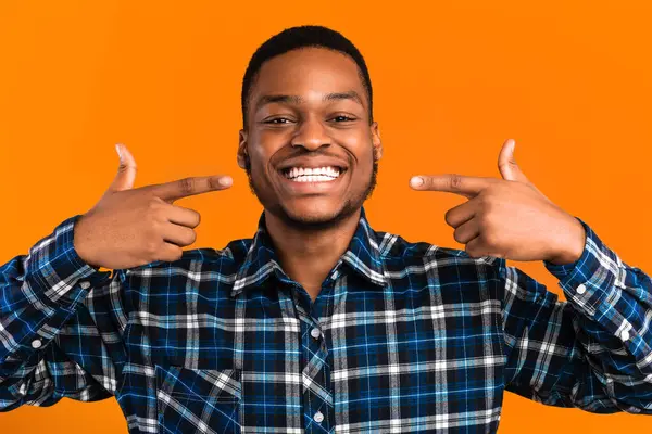 Gülümse. Neşeli siyah adam sarı arka planda kusursuz beyaz diş pozunu işaret ediyor. Stüdyo Çekimi