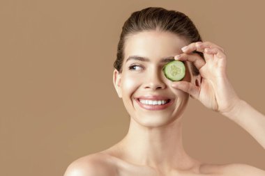 Doğal cilt temizleme konsepti. Bin yıllık çekici bir kadın gözlerinin önünde bir parça salatalık tutuyor ve kahverengi stüdyo arkaplanına gülümsüyor.