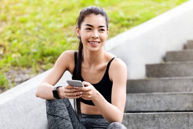Güzel genç Asyalı kadın dışarıda koştuktan sonra dinleniyor, akıllı telefonuyla şehir merdivenlerinde oturuyor.