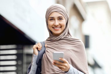 Sırt çantalı neşeli genç bir Arap kadın sokakta yürüyor, akıllı telefon kullanıyor, kopyalama alanı kullanıyor.