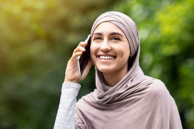 Neşeli Arap kadın parkta eğleniyor, çimenlerde oturuyor ve telefonda konuşuyor, boş alan.