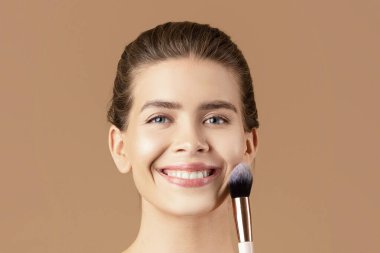 Cildi pürüzsüz, kahverengi stüdyo arka planında makyaj fırçası koleksiyonu olan mutlu genç bir kadının portresi. Süslü kozmetik ürünleri uygulayan, kameraya gülümseyen seksi milenyum kadını.