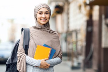 Müslüman kız üniversiteye gidiyor, elinde not defterleri ve sokakta kameraya gülümsüyor, eğitici bir konsept.