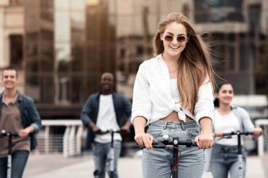Dört genç şehir merkezinde eğlenceli bir gezintiden zevk alıyor, düz bir caddede scooterlara biniyorlar. Modern binalar ve güneş ışığı altında süzülürken bir yoldaşlık duygusu sergiliyorlar..