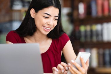 Kafeteryadaki dizüstü bilgisayarda çalışırken akıllı telefon kullanan, internete mesaj atan ya da gezen güzel Asyalı kadın.