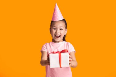 Tatilleri kutluyoruz. Beyaz tişörtlü, parti şapkalı kırmızı kurdeleli heyecanlı Asyalı çocuğun portresi sarı stüdyo arka planında izole bir şekilde duruyor.