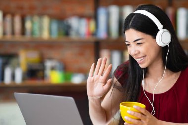 Video çağrısı. Kulaklıklı arkadaş canlısı Asyalı kız kafede dizüstü bilgisayar kullanıyor, kameraya el sallıyor, boş yer var.