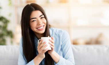 Gülümseyen Koreli kadın evdeki kanepede dinlenirken kahve içiyor, kameraya bakıyor, oturma odasında kanepede dinlenirken sıcak içkinin tadını çıkarıyor, kapalı bir yerde tatil yapıyor, yakın plan portre çekiyor, boş alan arıyor.