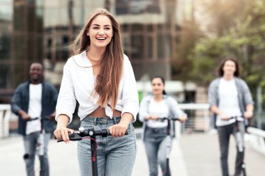 Anın tadını çıkarıyorum. Arka planda çetesiyle caddenin aşağısında stand-up scooter kullanan heyecanlı genç bir kız.